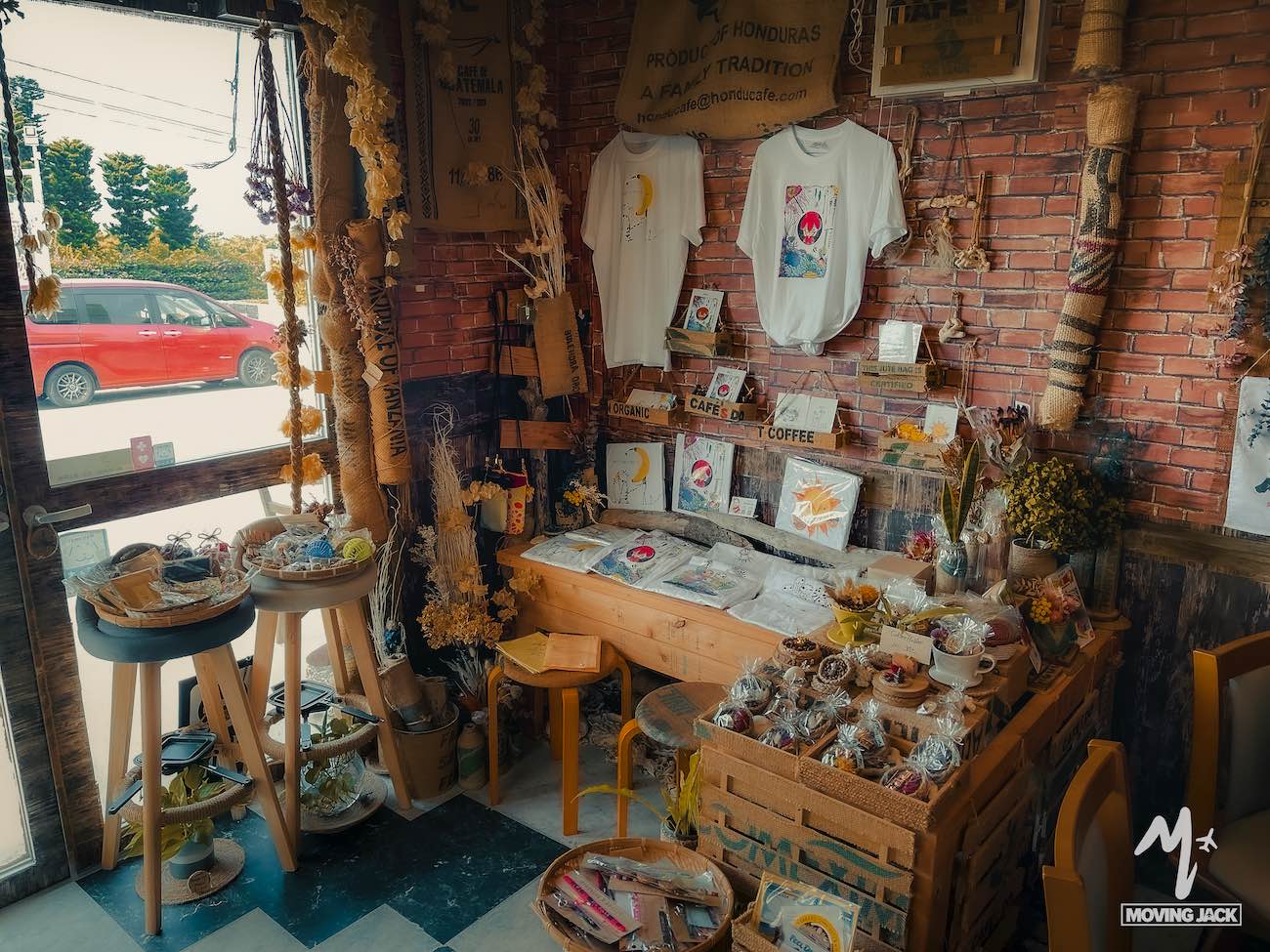 A cozy shop corner with wooden shelves displaying crafts, dried flowers, snacks, and art—an ideal stop on your okinawa itinerary. Sunlight streams in as two white graphic t-shirts hang on the brick wall above the displays. -copyright-moving-jack. Com