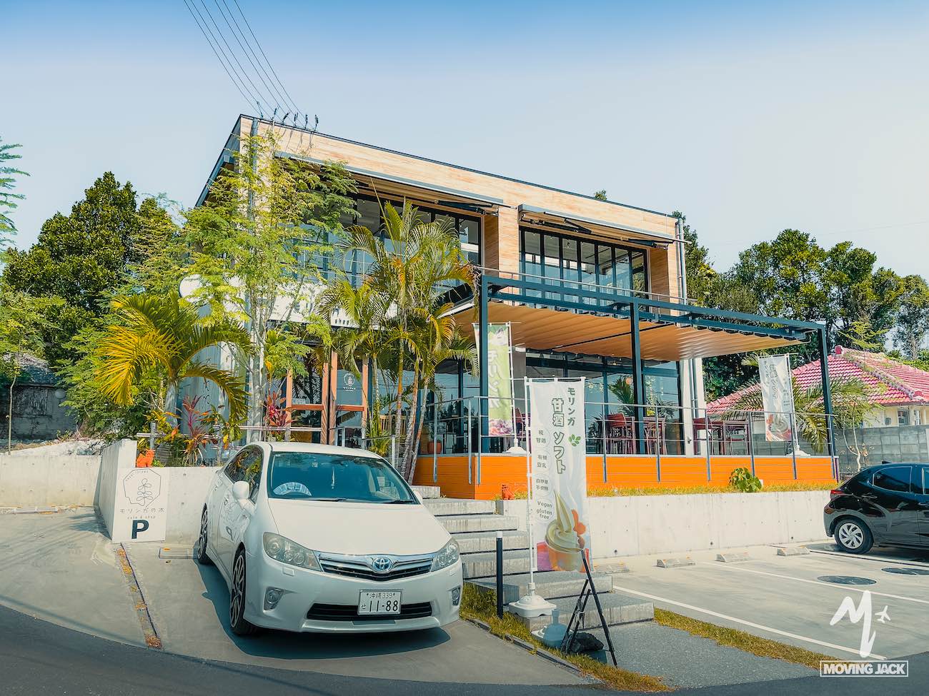 A modern, two-story café with large glass windows and wooden accents, surrounded by lush greenery—a perfect stop on your okinawa itinerary. A white car is parked in front, with banners and palm trees decorating the entrance on a sunny day. -copyright-moving-jack. Com