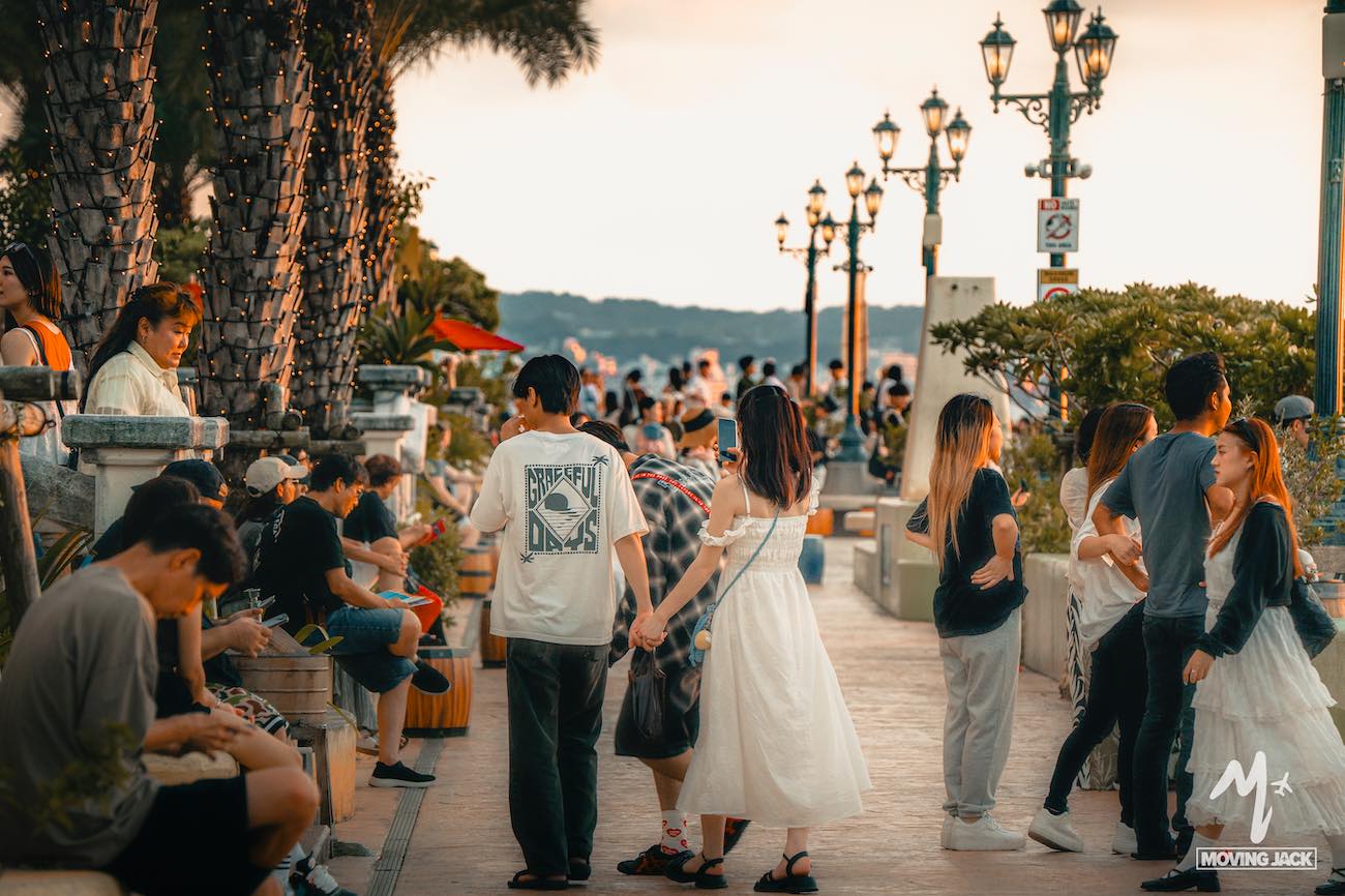 A lively outdoor scene at sunset shows people socializing along a scenic walkway lined with palm trees and lampposts—an inviting stop for any okinawa itinerary. A couple holds hands while groups relax and enjoy the tranquil atmosphere. -copyright-moving-jack. Com