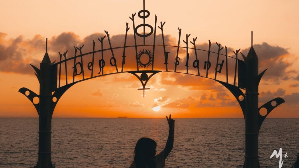26 okinawa tips first-time visitors need to know 1 A person sits beneath a decorative “depa island” sign, silhouetted against an orange sunset over the ocean—an iconic moment for any okinawa itinerary. Scattered clouds drift across the sky as they make a peace sign with their hand. -copyright-moving-jack. Com
