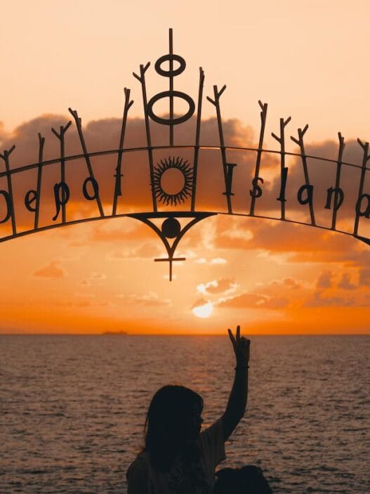 A person sits beneath a decorative “depa island” sign, silhouetted against an orange sunset over the ocean—an iconic moment for any okinawa itinerary. Scattered clouds drift across the sky as they make a peace sign with their hand. -copyright-moving-jack. Com