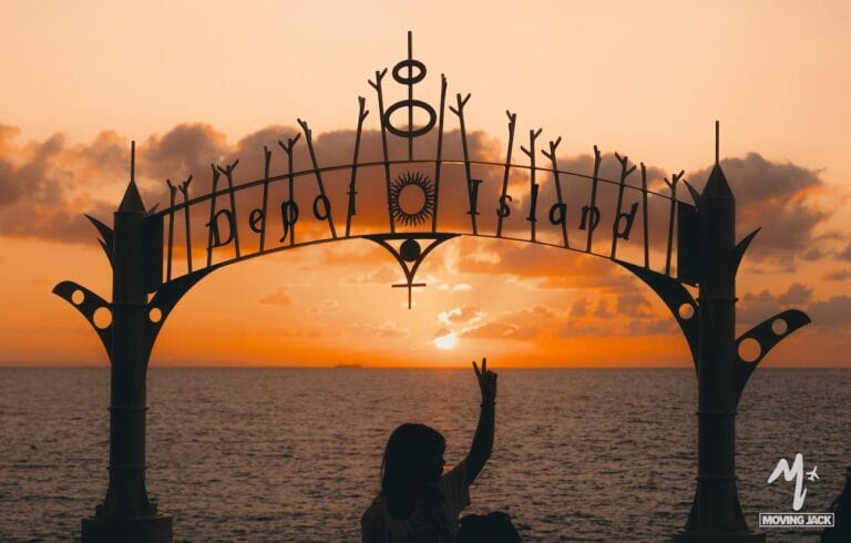 Een persoon zit onder een decoratief bord met "Depa Island", afgetekend tegen een oranje zonsondergang boven de oceaan – een iconisch moment voor elke Okinawa-reis. Verspreide wolken drijven door de lucht terwijl ze met hun hand een vredesgebaar maken. -copyright-moving-jack.com