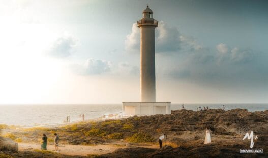 A tall lighthouse stands on rocky terrain by the sea under a partly cloudy sky—a perfect stop for any okinawa itinerary. Several people are scattered around the rocks, taking photos as sunlight filters through the clouds and the ocean glimmers in the background. -copyright-moving-jack. Com
