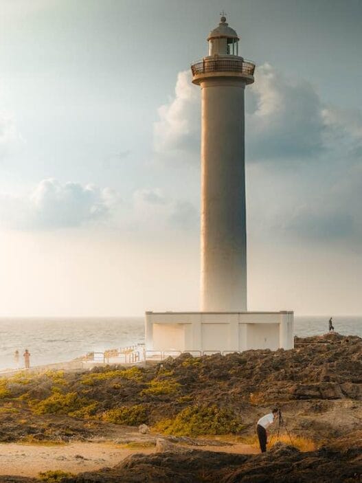 Okinawa itinerary: best 3, 5, or 7 days of relaxing and fun 53 A tall lighthouse stands on rocky terrain by the sea under a partly cloudy sky—a perfect stop for any okinawa itinerary. Several people are scattered around the rocks, taking photos as sunlight filters through the clouds and the ocean glimmers in the background. -copyright-moving-jack. Com