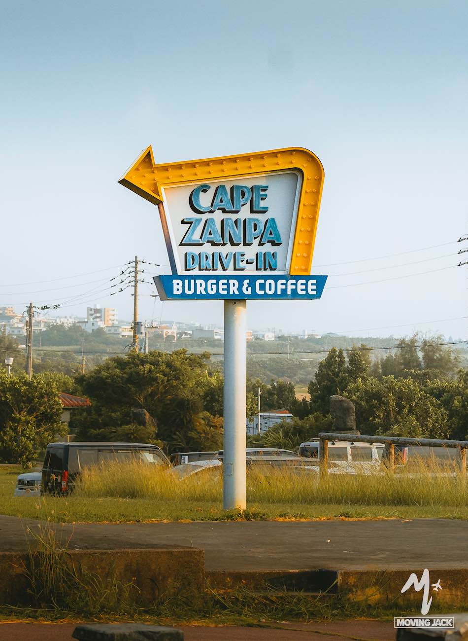 26 okinawa tips first-time visitors need to know 4 A retro-style sign with a yellow arrow reads "cape zanpa drive-in burger & coffee. " perfect for your okinawa itinerary, the sign stands beside a grassy field with parked cars, trees, and power lines in the background. -copyright-moving-jack. Com