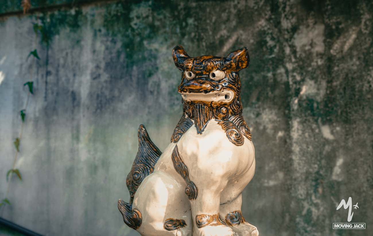 26 okinawa tips first-time visitors need to know 5 A ceramic shisa lion-dog statue with brown detailing sits in front of a weathered, mossy wall. The statue has a fierce expression and detailed fur textures. The moving jack logo appears in the bottom right corner. -copyright-moving-jack. Com