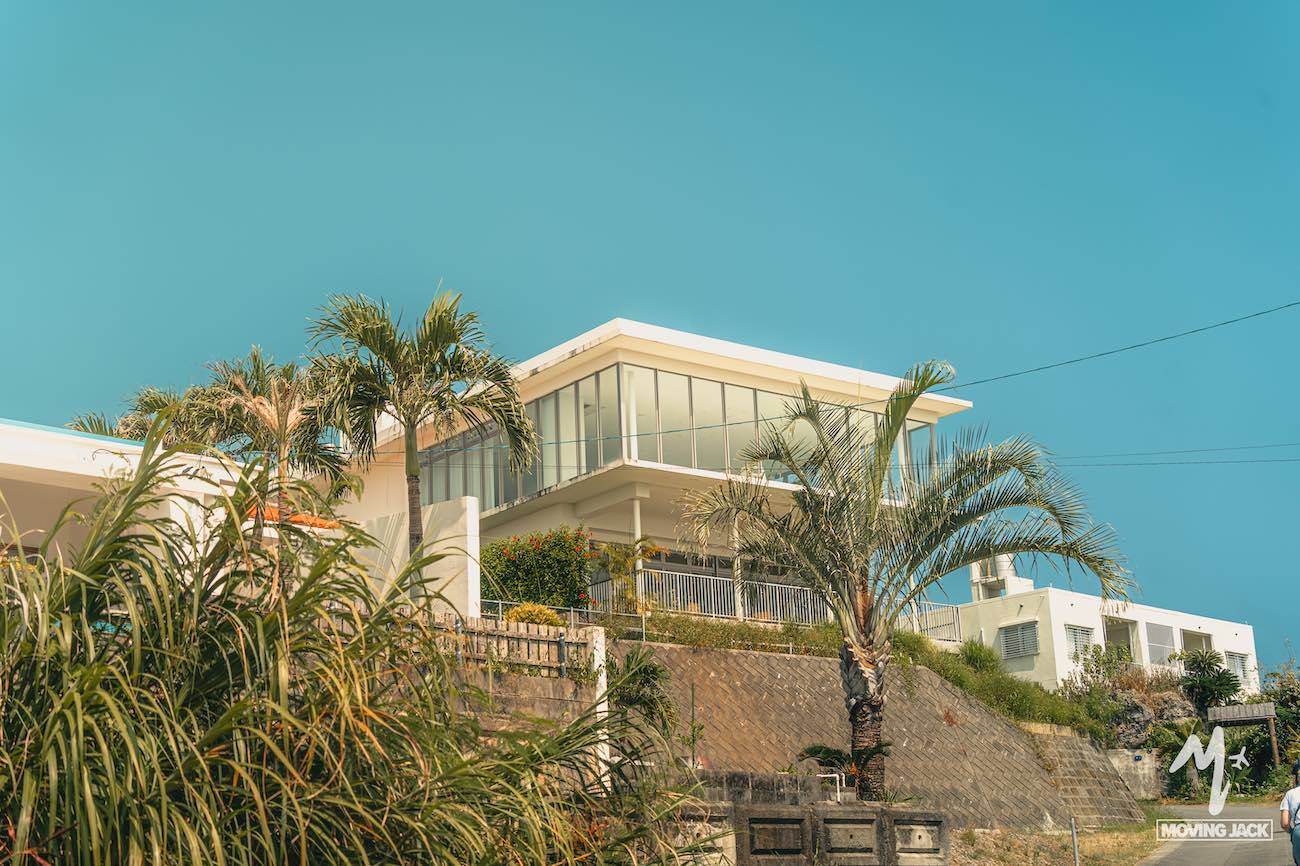 26 okinawa tips first-time visitors need to know 20 A modern white house with large glass windows is surrounded by palm trees and greenery under a clear blue sky. The building sits on a stone terrace, giving it an elevated appearance. -copyright-moving-jack. Com