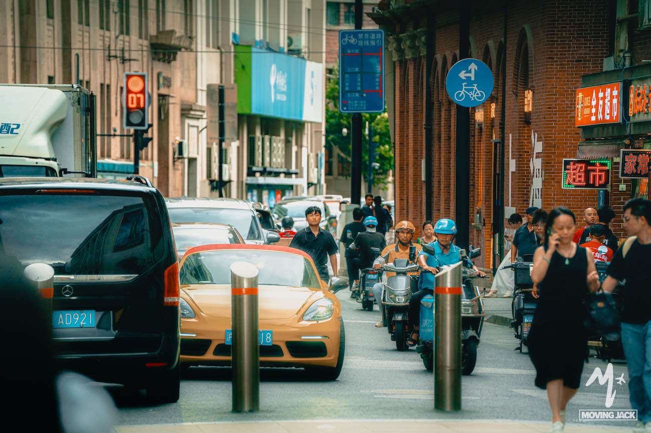 Een drukke stadsstraat met auto's, scooters en voetgangers – klassieke taferelen die je doen afvragen: is Shanghai een bezoek waard? Het levendige stadsleven ontvouwt zich terwijl mensen tussen wachtende voertuigen doorsteken en winkels de drukke trottoirs sieren. -Copyright-moving-jack.com