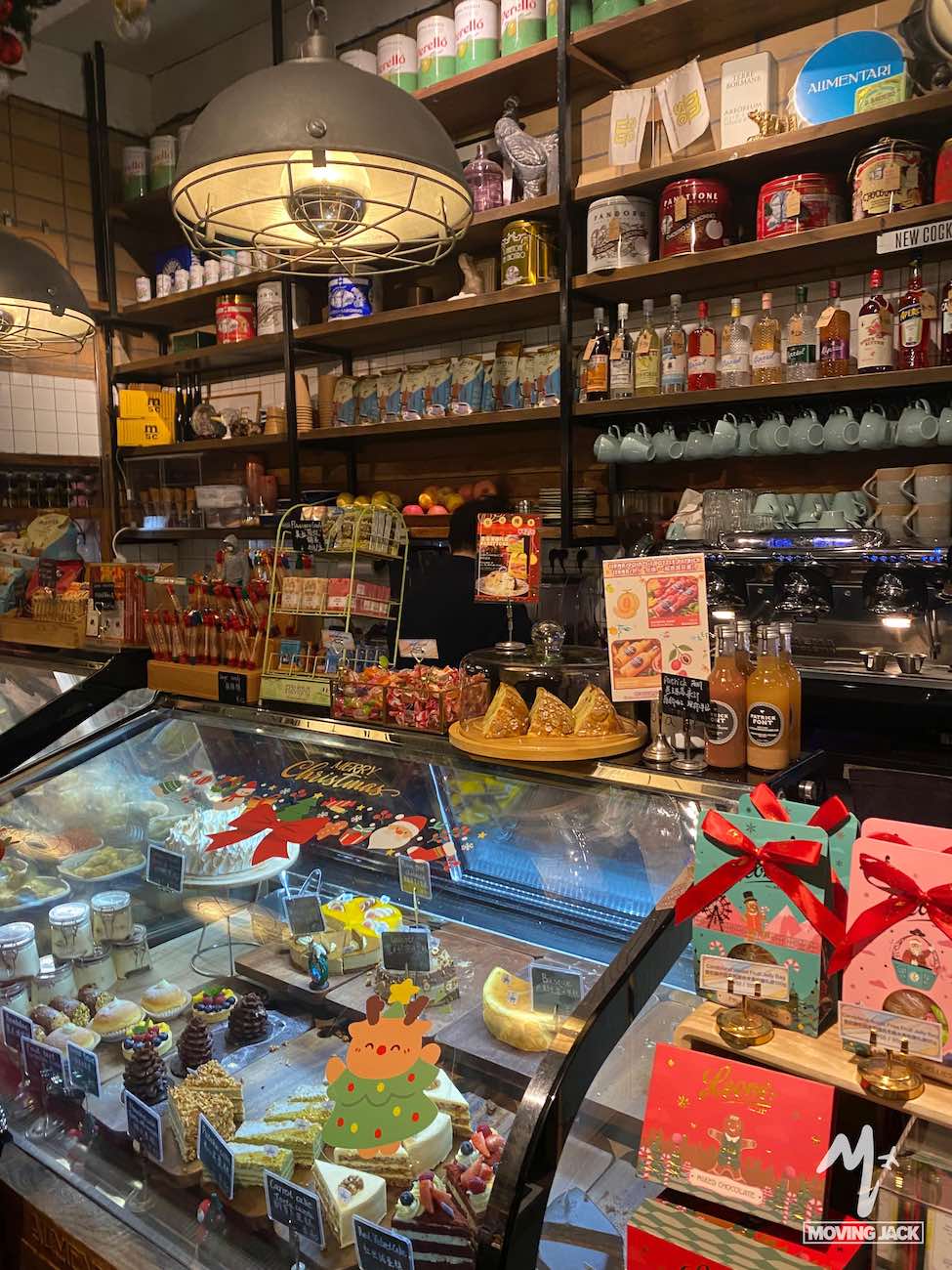 Een knusse delicatessenwinkel met schappen vol kleurrijke blikken, flessen en mokken. In een glazen vitrine liggen diverse kazen, gebakjes en desserts, met kerstversieringen en geschenkdozen op de voorgrond. -Copyright-moving-jack.com