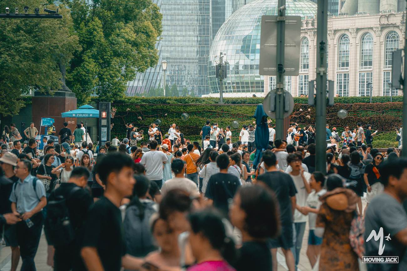 A large crowd of people gathers outdoors in front of lush greenery and modern glass buildings, some taking photos and others walking, on a sunny day in an urban setting. -Copyright-moving-jack.com