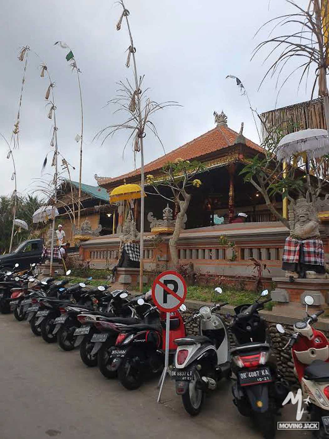 Een rij geparkeerde scooters staat langs de straat voor een traditioneel Balinees gebouw met sierlijke versieringen, geruite doeken rond beelden, hoge bamboepalen en een 'verboden te parkeren'-bord op de voorgrond. -copyright-moving-jack.com