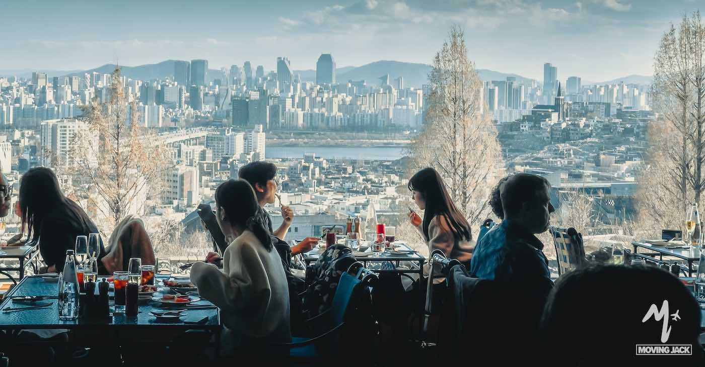 People dine at a restaurant with large windows overlooking a city skyline, tall buildings, and a river in the background. Daylight illuminates the scene, with trees framing the urban view. -copyright-moving-jack. Com