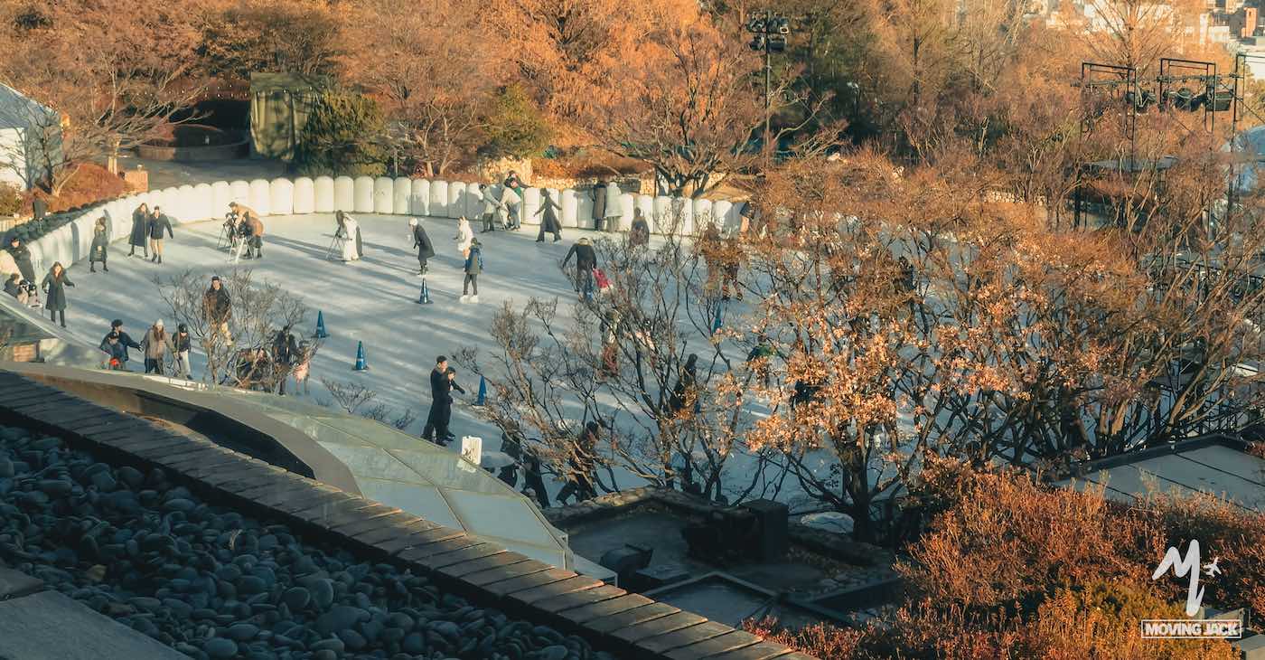 People ice skating on an outdoor rink surrounded by autumn trees with orange leaves. The scene is bathed in warm sunlight, and some visitors stand or watch around the edges of the rink. -copyright-moving-jack. Com