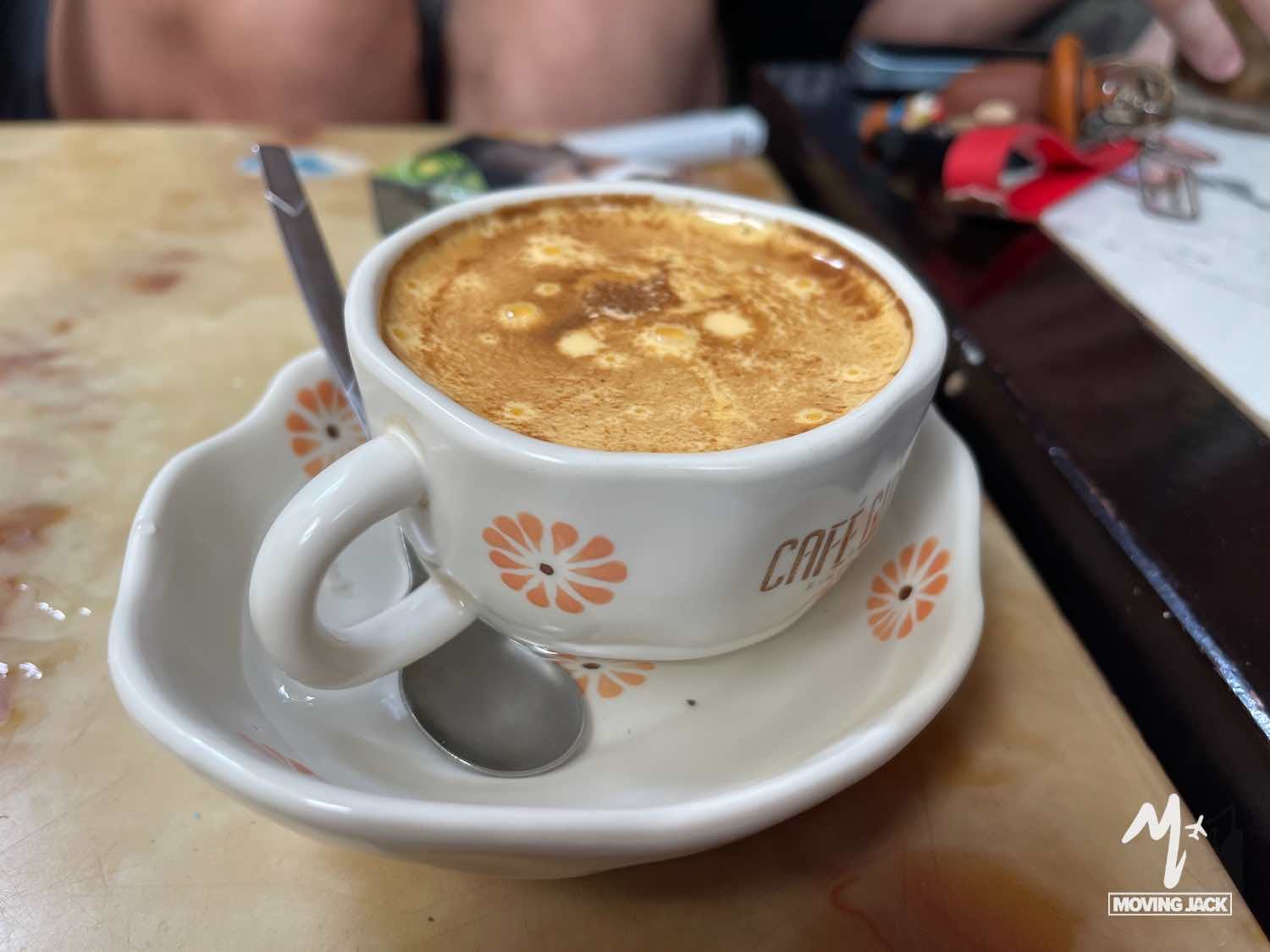 Een kopje schuimige Vietnamese eierkoffie staat op een schoteltje met een lepeltje op een beige tafel. Het kopje is versierd met oranje bloemmotieven. Sleutels en andere voorwerpen zijn wazig op de achtergrond. -Copyright-moving-jack.com