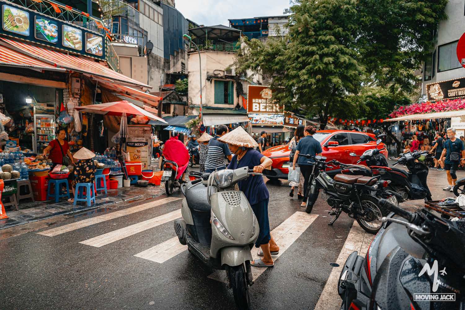Een drukke straatmarkt in Vietnam met mensen die kegelvormige hoeden dragen, motoren en auto's, omringd door eetkraampjes, winkels en kleurrijke paraplu's op een levendige, regenachtige dag. -Copyright-moving-jack.com