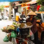 A street vendor wearing a conical hat stands beside a bicycle laden with fruits on a rainy city street, next to an orange car and under a blue umbrella. Other vehicles and people are visible in the background. -copyright-moving-jack. Com