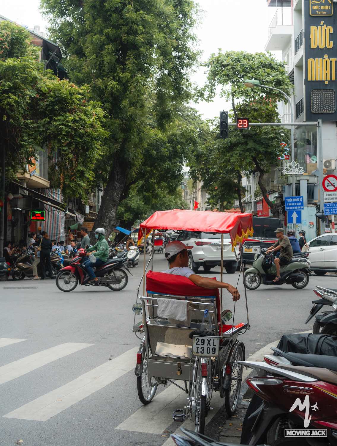 Een man met een wit shirt en een pet zit in een fietstaxi op een druk kruispunt in de stad. Motorfietsen en voetgangers passeren, en een verkeerslicht staat op rood. Bomen en hoge gebouwen flankeren de straat. -Copyright-moving-jack.com