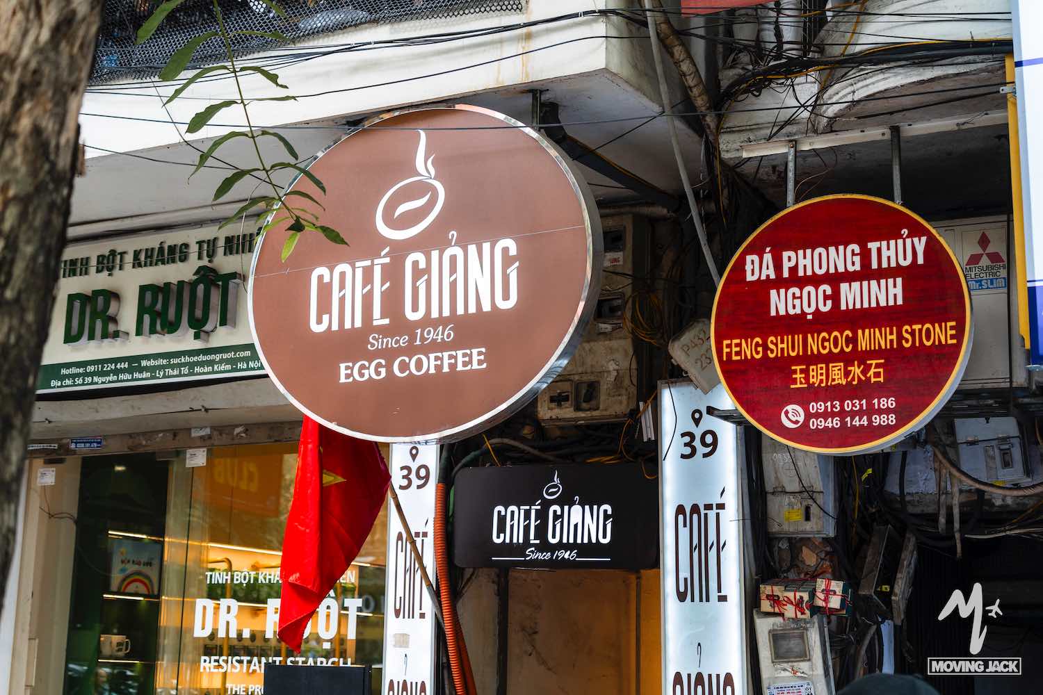 Een straatbeeld toont het ronde bruine bord van "Café Giảng Egg Coffee" en een rood bord van "Phong Thủy Ngọc Minh" tussen winkelgevels en diverse andere uithangborden in een drukke stedelijke omgeving. -Copyright-moving-jack.com