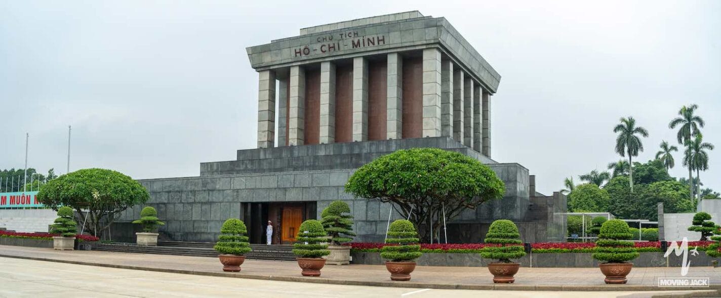 A wide view of Ho Chi Minh Mausoleum in Hanoi, Vietnam, a must-see among things to do in Hanoi, featuring a gray stone building with columns, surrounded by neatly trimmed trees and red flowers under a cloudy sky. -Copyright-moving-jack.com