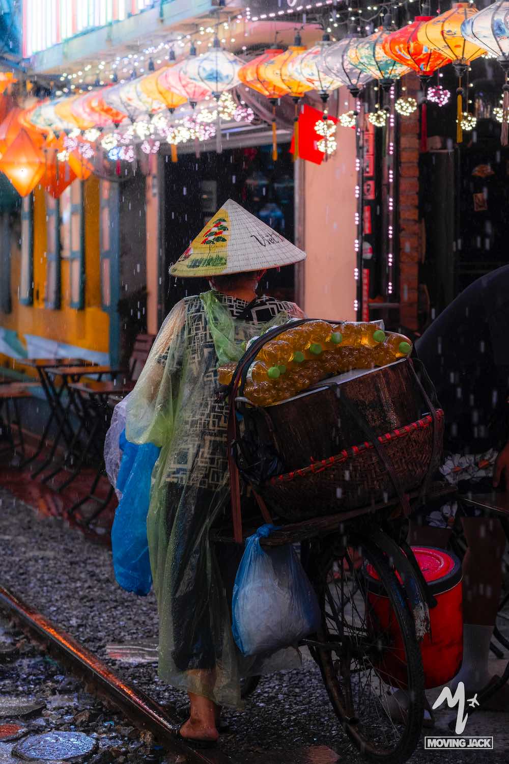 Een persoon met een kegelvormige hoed en een regenponcho loopt naast een fiets beladen met manden vol goederen op een regenachtige straat, verlicht door kleurrijke lantaarns en lampen. -Copyright-moving-jack.com