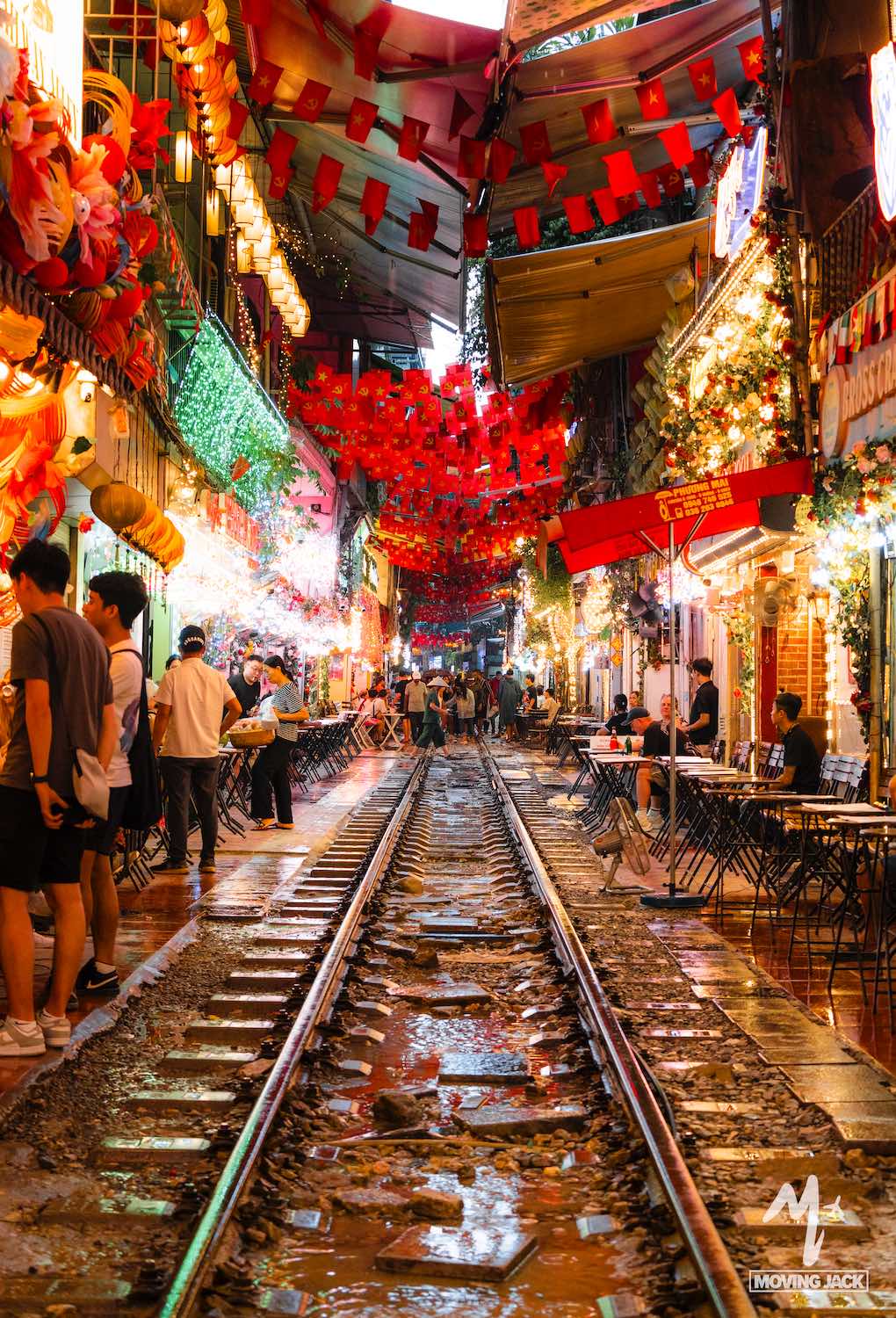 A lively street scene in Hanoi with railway tracks running down the center, lined with colorful lanterns, bright lights, outdoor seating, and people dining in a festive atmosphere—one of the must-see things to do in Hanoi. -Copyright-moving-jack.com