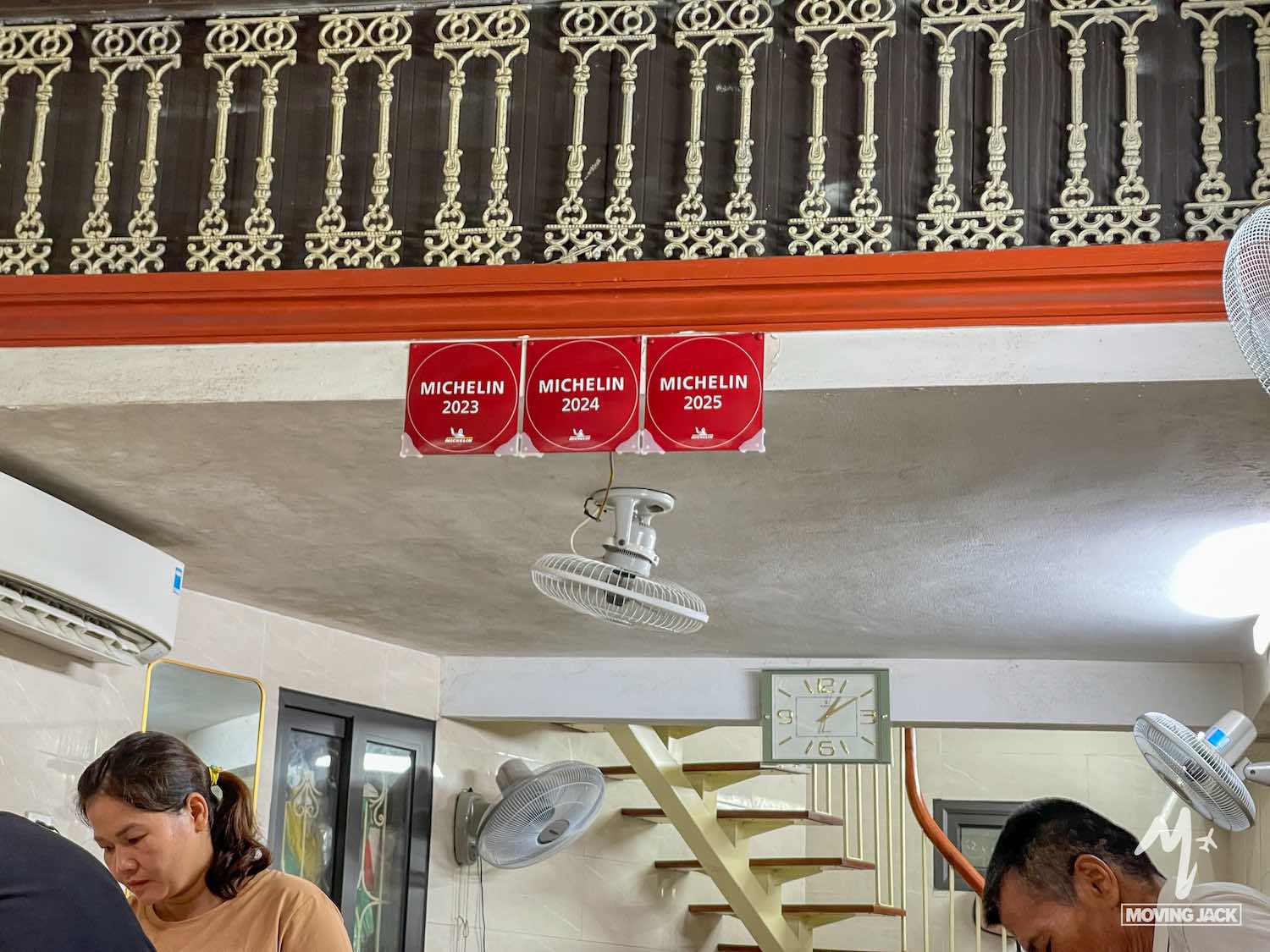 Een restaurantinterieur met drie rode Michelin-borden voor 2023, 2024 en 2025 aan de muur, een sierlijke zwarte balustrade erboven, een witte wandventilator, een trap en twee mensen die beneden aan het werk zijn. -Copyright-moving-jack.com