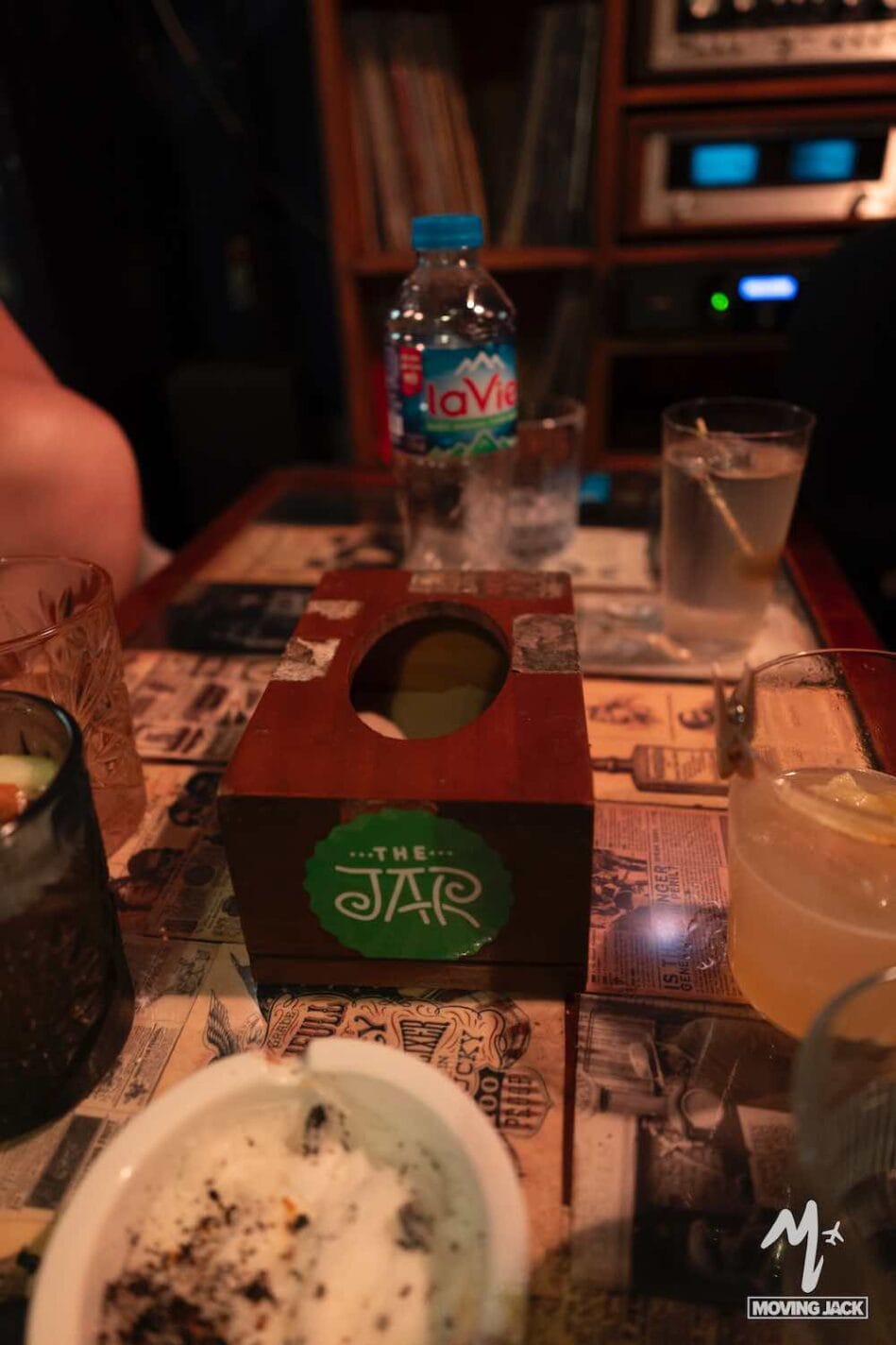 Een houten tissuebox met een groene "The JAR"-sticker staat op een tafel met drankjes, een waterfles en een asbak – perfect voor wie op zoek is naar unieke dingen om te doen in Hanoi, te midden van vintage decor en retro audioapparatuur. -Copyright-moving-jack.com