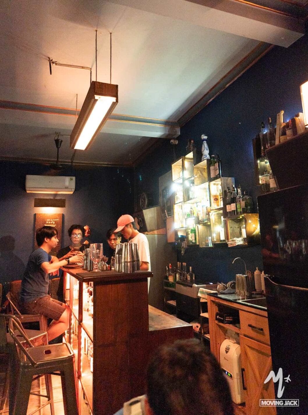Vier mensen zitten aan een schemerig verlichte bar met planken vol flessen aan de muur. Een barman serveert drankjes terwijl drie gasten met elkaar praten. De ruimte heeft donkerblauwe muren en een gezellige, ongedwongen sfeer. -Copyright-moving-jack.com