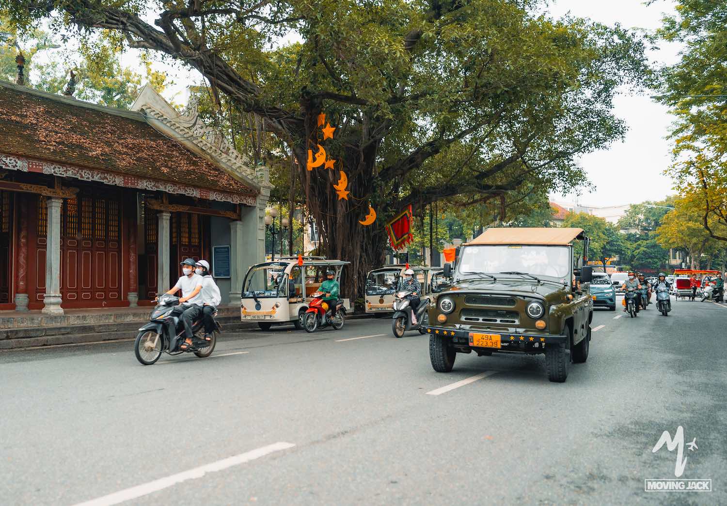 Een straatbeeld toont motoren, auto's en tuk-tuks die langs een traditioneel gebouw en een grote boom met oranje lantaarns rijden, in wat een bruisende Aziatische stad lijkt te zijn. -Copyright-moving-jack.com