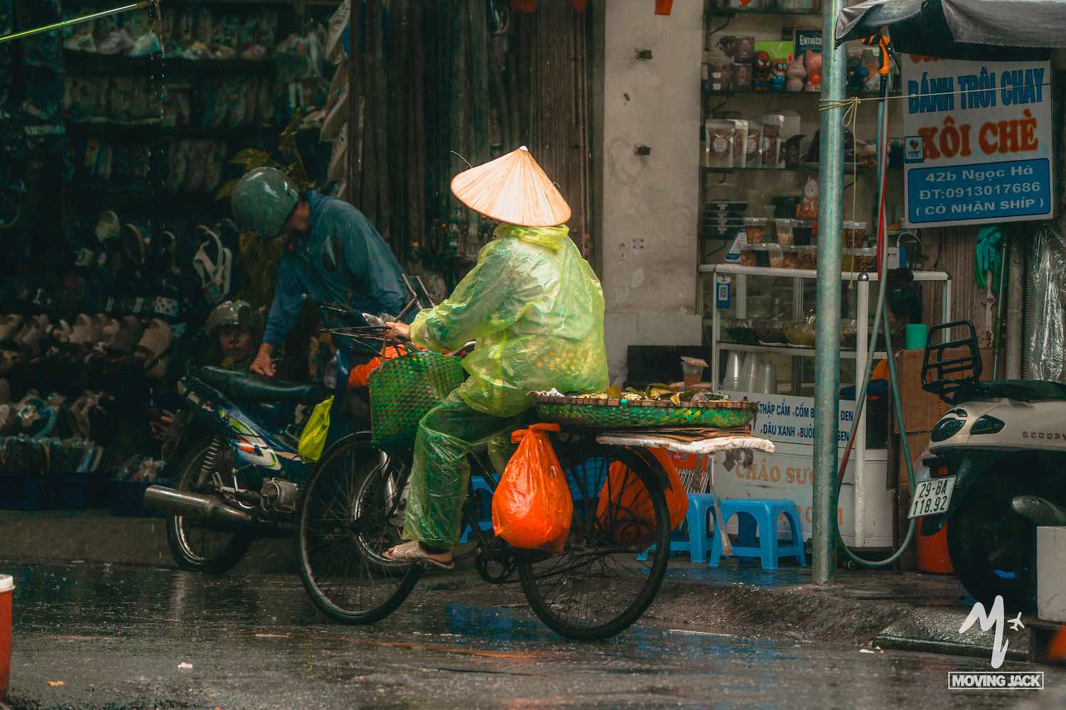 Een persoon met een kegelvormige hoed en een groene regenjas rijdt op een fiets beladen met goederen door een regenachtige straat in Vietnam, langs winkels en een andere persoon op een motorfiets. -Copyright-moving-jack.com