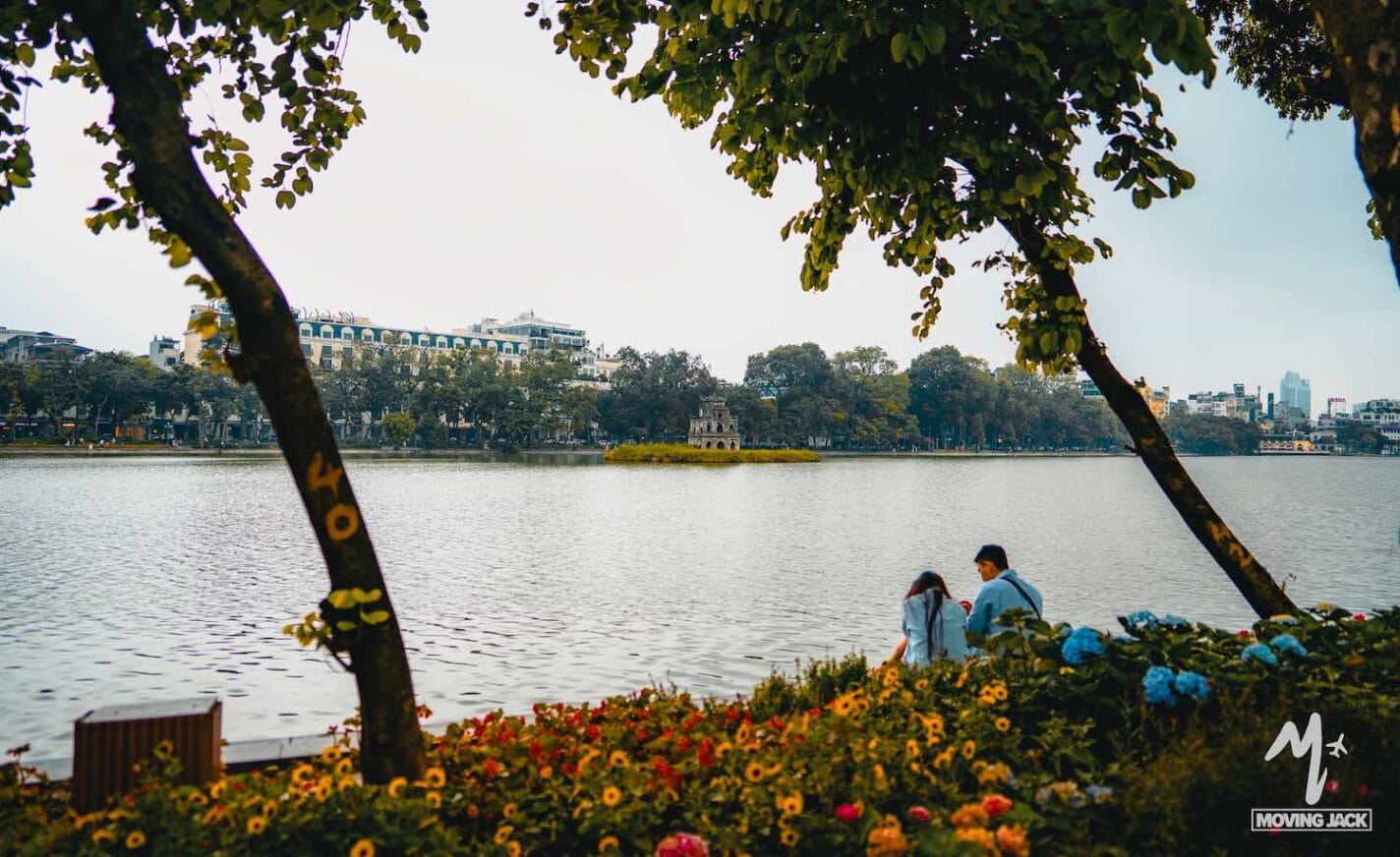 Two people sit by a lake surrounded by colorful flowers and trees, with buildings and a small tower visible across the water under a bright sky—a scene that captures the charm of things to do in Hanoi. -Copyright-moving-jack.com
