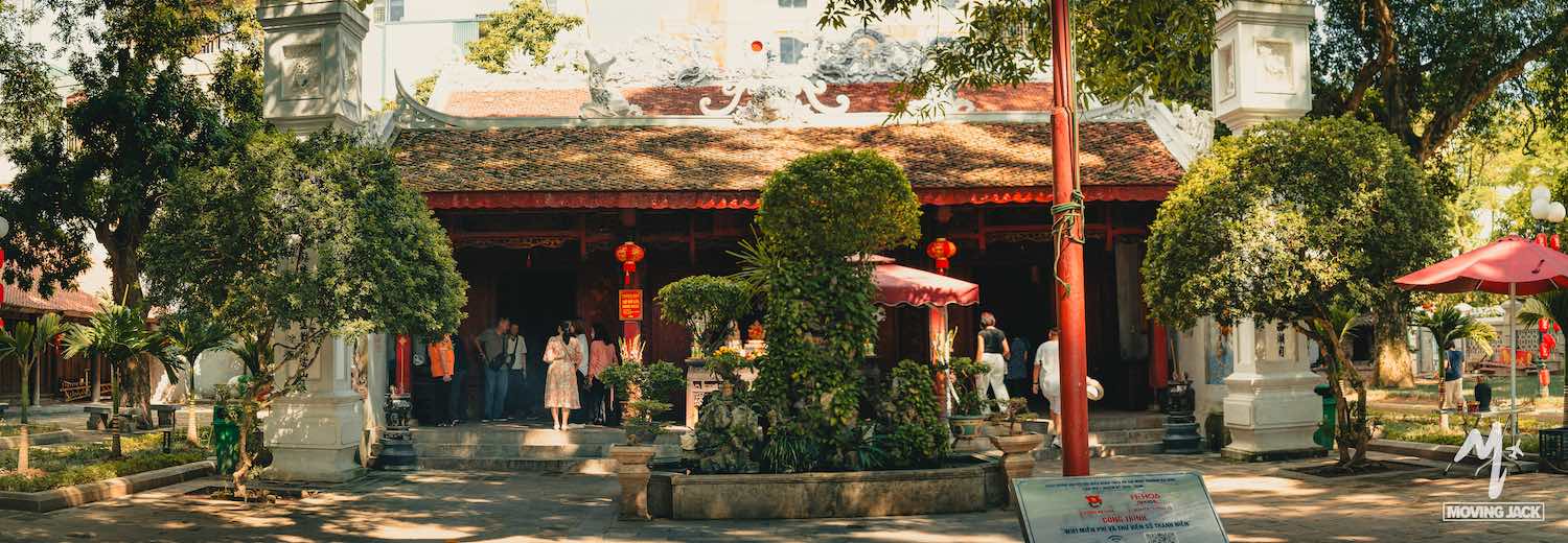 Een groep mensen staat voor een traditionele Vietnamese tempel met een sierlijk rood dak, stenen beelden, rode lantaarns en weelderige bomen – een absolute aanrader voor een zonnige dag in Hanoi. -Copyright-moving-jack.com