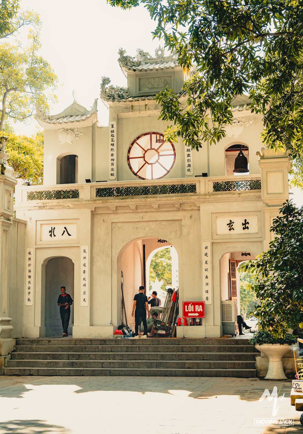 Een traditionele Vietnamese poort met witte muren, gedetailleerde dakversieringen en Chinese karakters verwelkomt bezoekers. Beschut door bomen is het een populaire stop voor wie Hanoi wil verkennen. Een rood bord met "LỐI RA" (uitgang) markeert de ingang. -Copyright-moving-jack.com