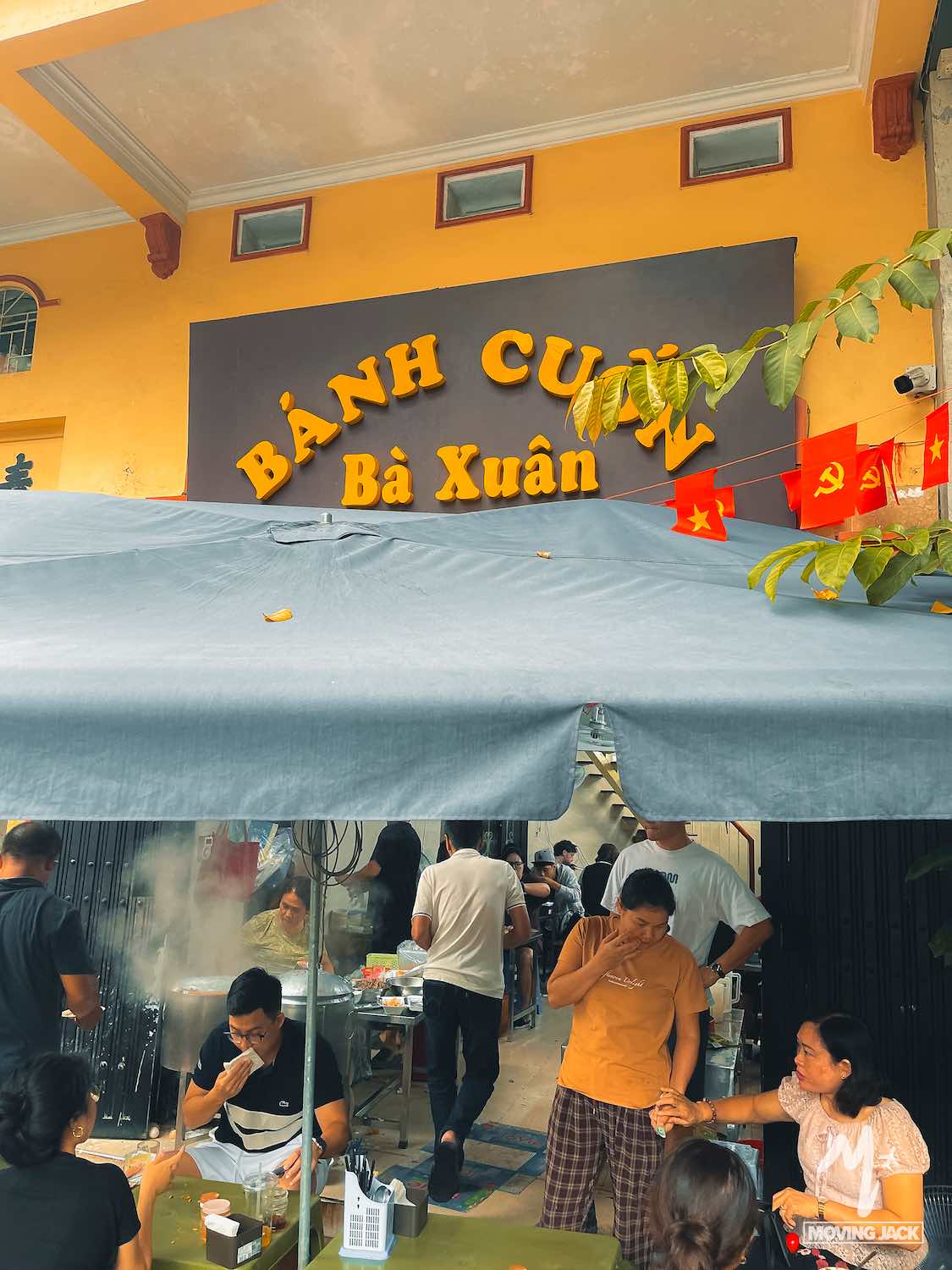 Mensen verzamelen zich om te eten buiten een populaire gele winkel met een groot bord waarop "Bánh Cuốn Bà Xuân" staat. Deze levendige plek, die vaak wordt genoemd als een van de beste dingen om te doen in Hanoi, biedt dampende gerechten die worden bereid onder een blauwe luifel en rode Vietnamese vlaggen. -Copyright-moving-jack.com