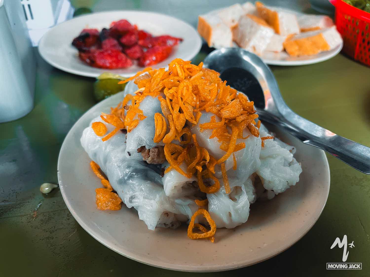Een bord Vietnamese gestoomde rijstrolletjes met gebakken sjalotten, met op de achtergrond borden gegrild vlees en tofu op een groene tafel – een gerecht dat je absoluut moet proberen en een van de hoogtepunten van een bezoek aan Hanoi. -Copyright-moving-jack.com