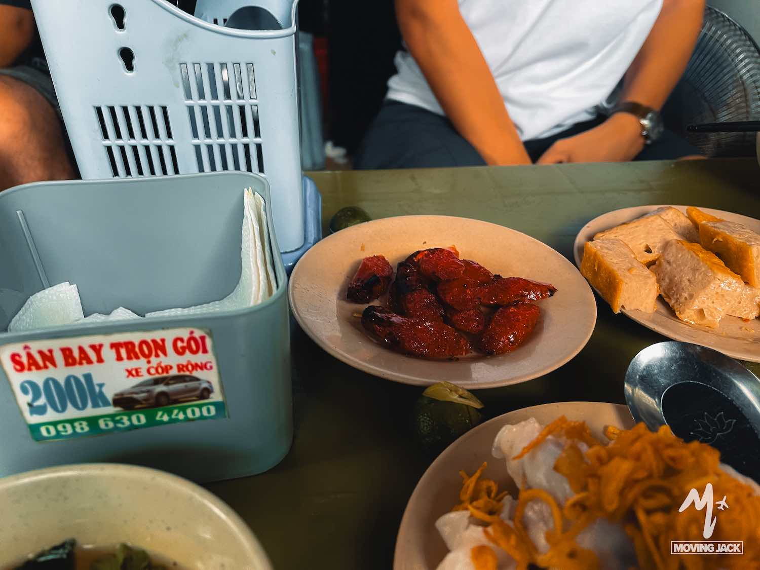Een tafel vol borden met Vietnamees eten, waaronder gegrild vlees, tofu en garnituren – perfect voor wie op zoek is naar leuke activiteiten in Hanoi. Op de achtergrond zit iemand met de armen over elkaar; een grijze plastic opbergbak en een bord met Vietnamese tekst zijn ook zichtbaar. -Copyright-moving-jack.com
