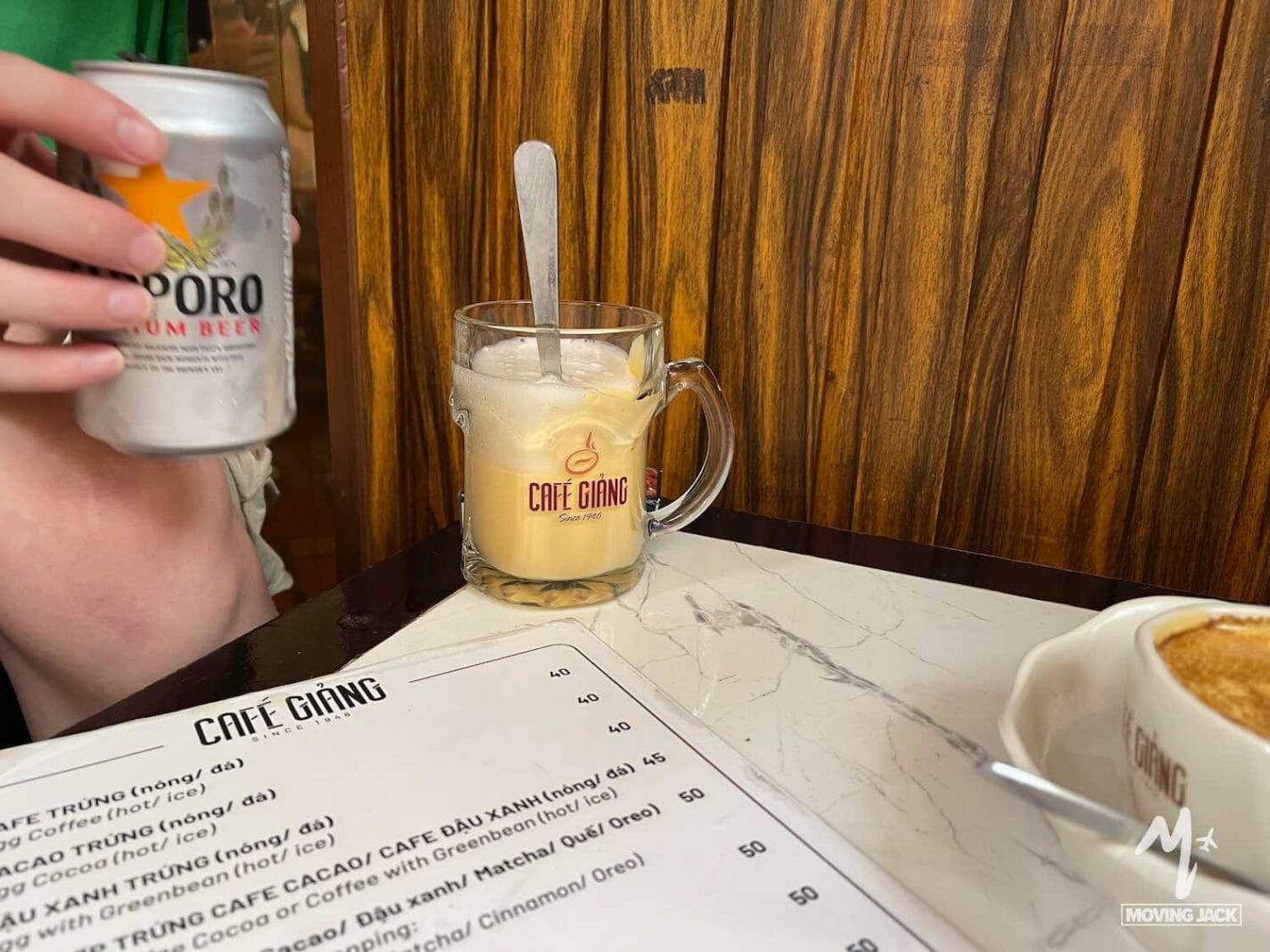 A hand holds a can of Sapporo beer beside a glass mug of egg coffee with a spoon at Café Giảng—one of the unique things to do in Hanoi. A menu and part of a coffee cup sit on a marble table, set against wood paneling. -Copyright-moving-jack.com