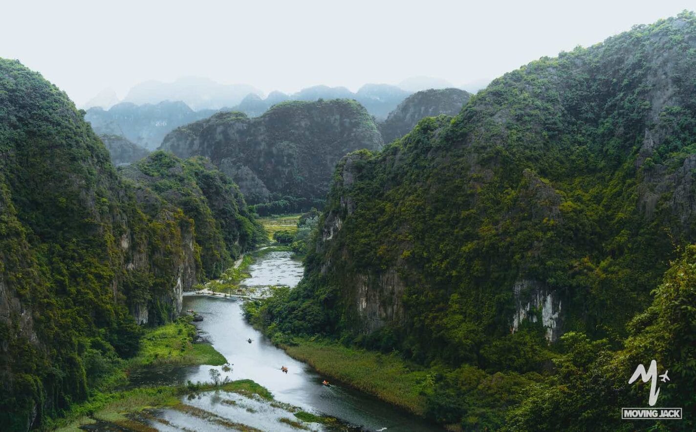 A lush, green river valley winds through towering, forested limestone mountains under a hazy sky, with small boats visible on the water below—one of the must-see Hanoi attractions for those seeking unique things to do in Hanoi. -Copyright-moving-jack.com