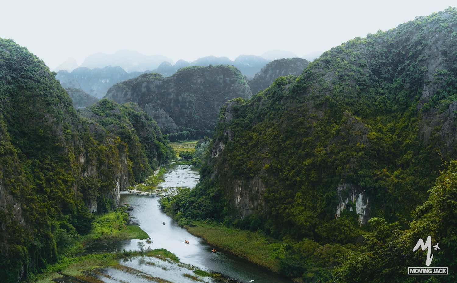 Een kronkelende rivier stroomt door een weelderig groene vallei, omgeven door hoge, rotsachtige bergen bedekt met vegetatie – die doen denken aan de schilderachtige landschappen die je in Hanoi kunt vinden – met kleine bootjes die op het mistige water drijven. -Copyright-moving-jack.com