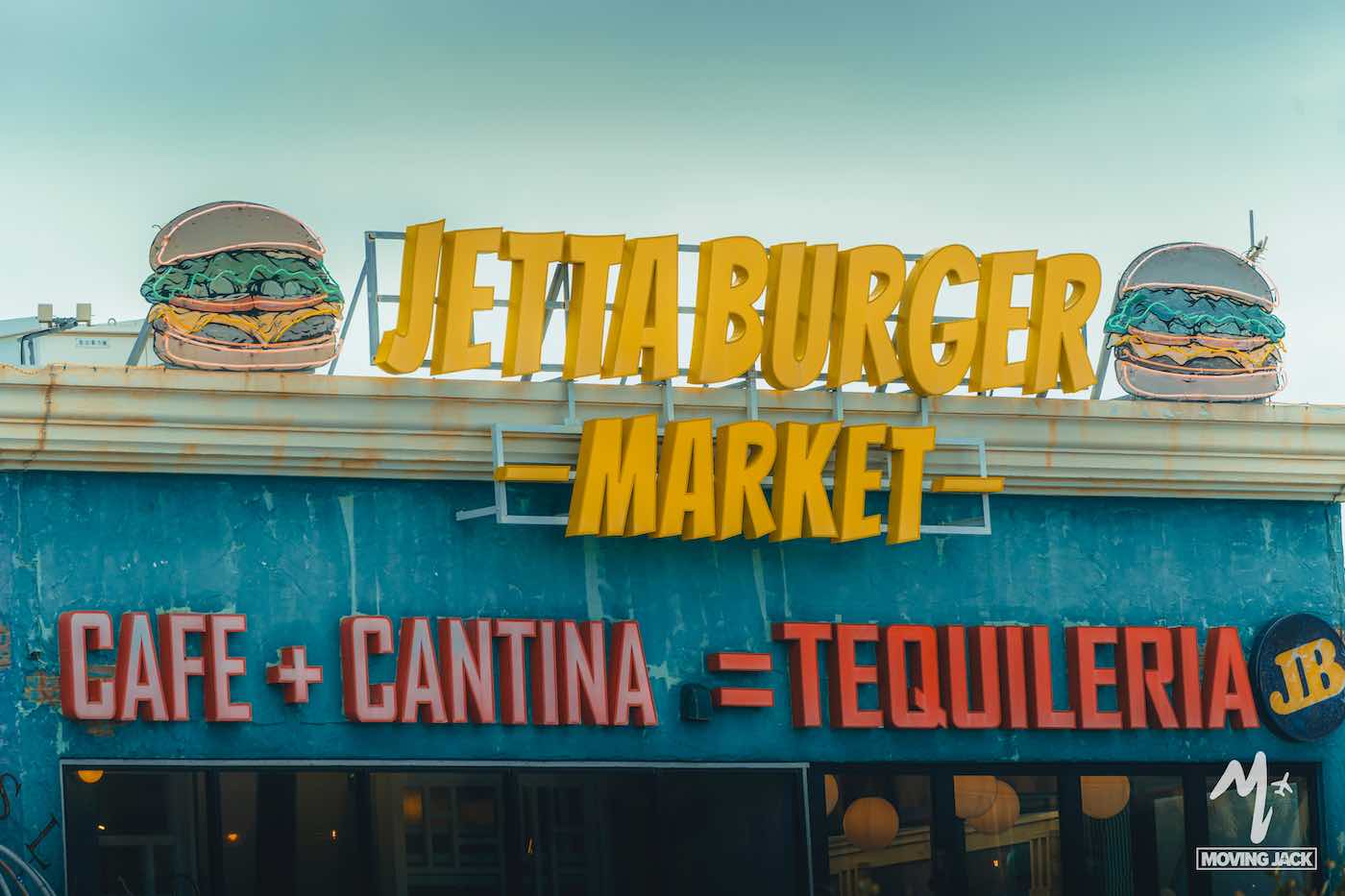 Op een restaurantbord staat "Jettaburger Market" met aan weerszijden afbeeldingen van hamburgers. Daaronder hangt een neonbord met de tekst "Café + Cantina = Tequileria" op een blauwe muur. Het Moving Jack-logo bevindt zich in de rechterbenedenhoek. -copyright-moving-jack.com