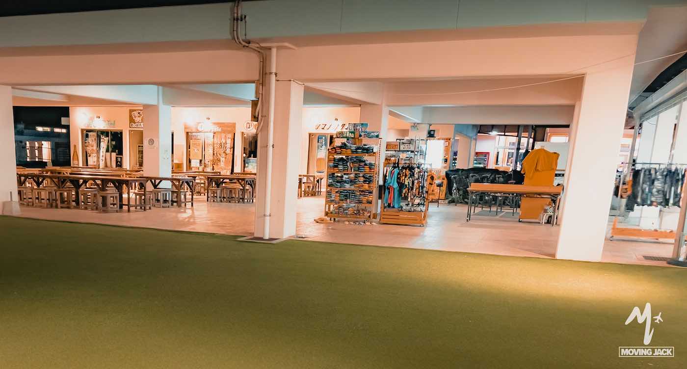 Nachtopname van een goed verlichte openluchtmarkt of foodcourt met houten tafels en stoelen, kledingrekken en diverse winkels. De sfeer oogt rustig en uitnodigend. -copyright-moving-jack.com