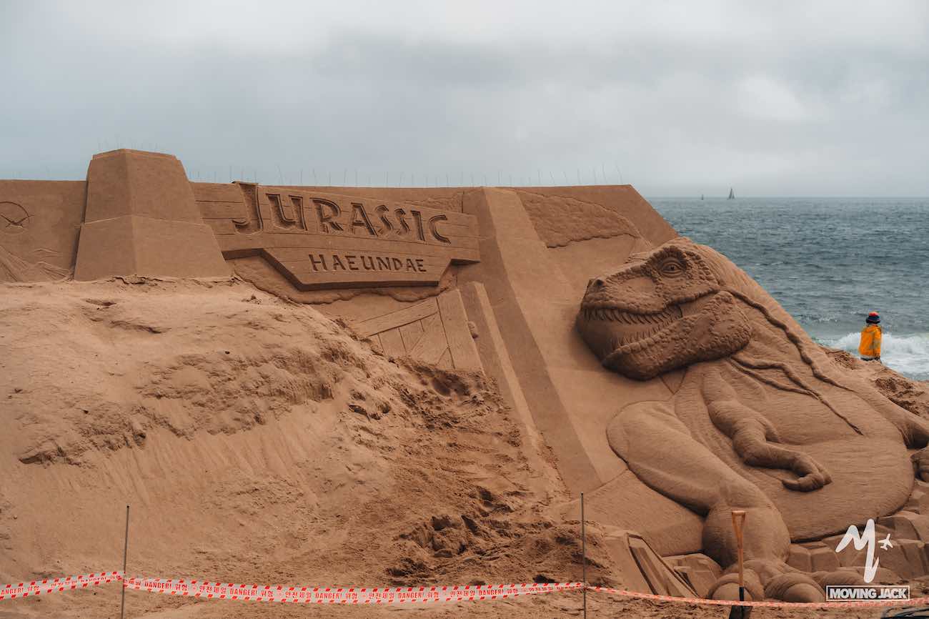 Large sand sculpture of a dinosaur with "JURASSIC HAEUNDAE" carved above, beside the ocean—just one reason why many say Busan is worth visiting.
