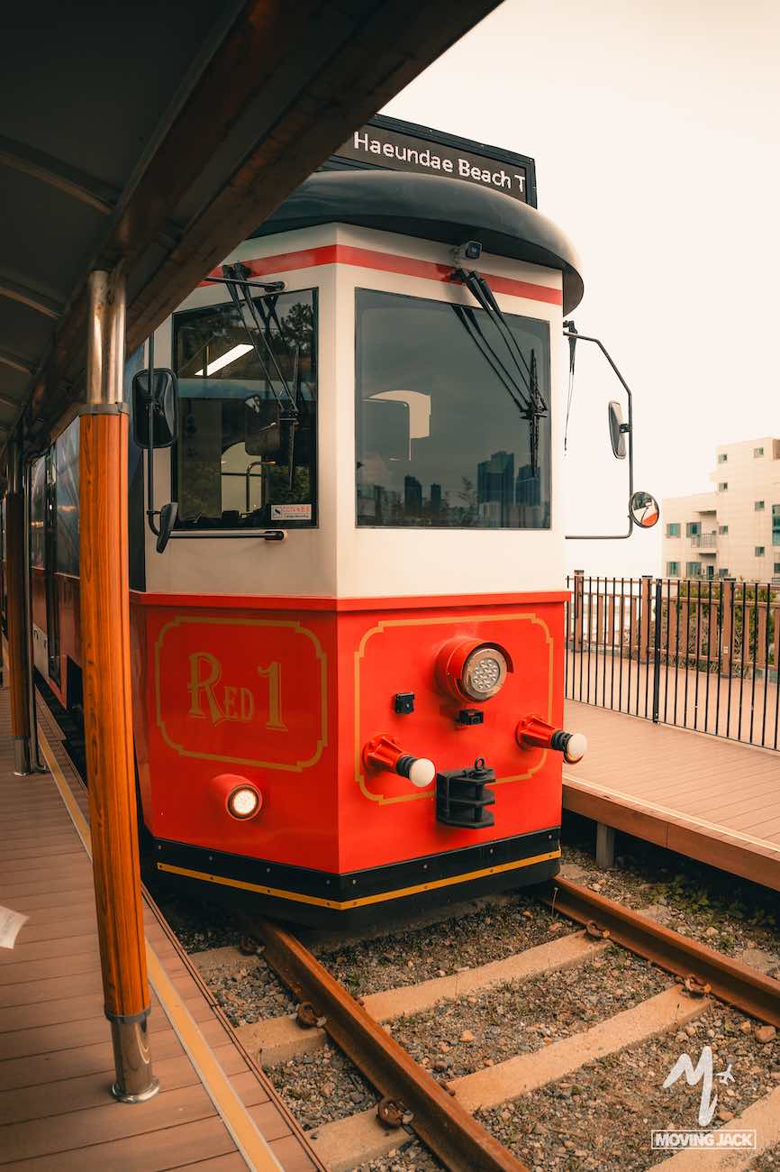 Red vintage tram labeled "Red 1" at a station, with a "Haeundae Beach" sign on top—a scene that answers is Busan worth visiting with its unique charm.