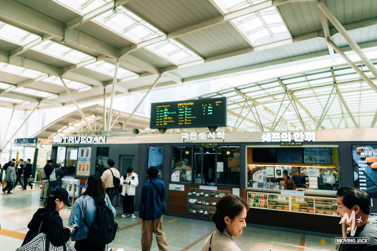 People walking inside a modern train station with signs and a digital departures board in Korean, offering a glimpse into daily life and raising the question: is Busan worth visiting for its vibrant urban atmosphere?.