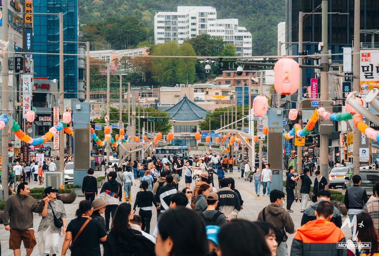 A busy city street with crowds, colorful lanterns, and buildings in the background captures the vibrant energy that makes you wonder: is Busan worth visiting?.