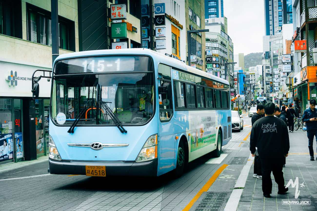 A light blue city bus numbered 145-1 drives down a busy street lined with shops and signs, offering a glimpse of daily life that makes you wonder— is Busan worth visiting?.