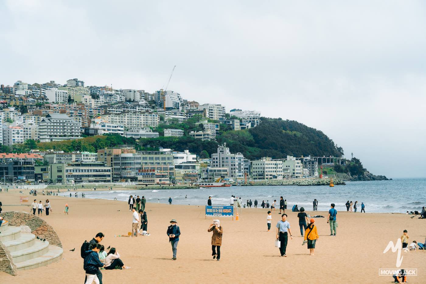 People walking and relaxing on a sandy beach with a cityscape and hills in the background under cloudy skies make you wonder: is Busan worth visiting? This picturesque scene shows why so many travelers say yes.
