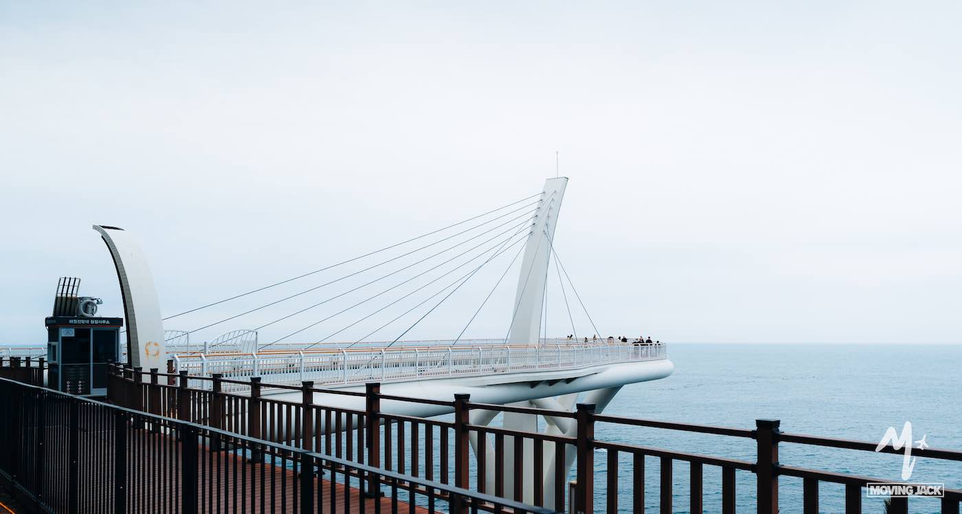 A modern observation deck with cables extends over the ocean, where people stand at the edge under a cloudy sky—scenes like this make you wonder, is Busan worth visiting? Absolutely.