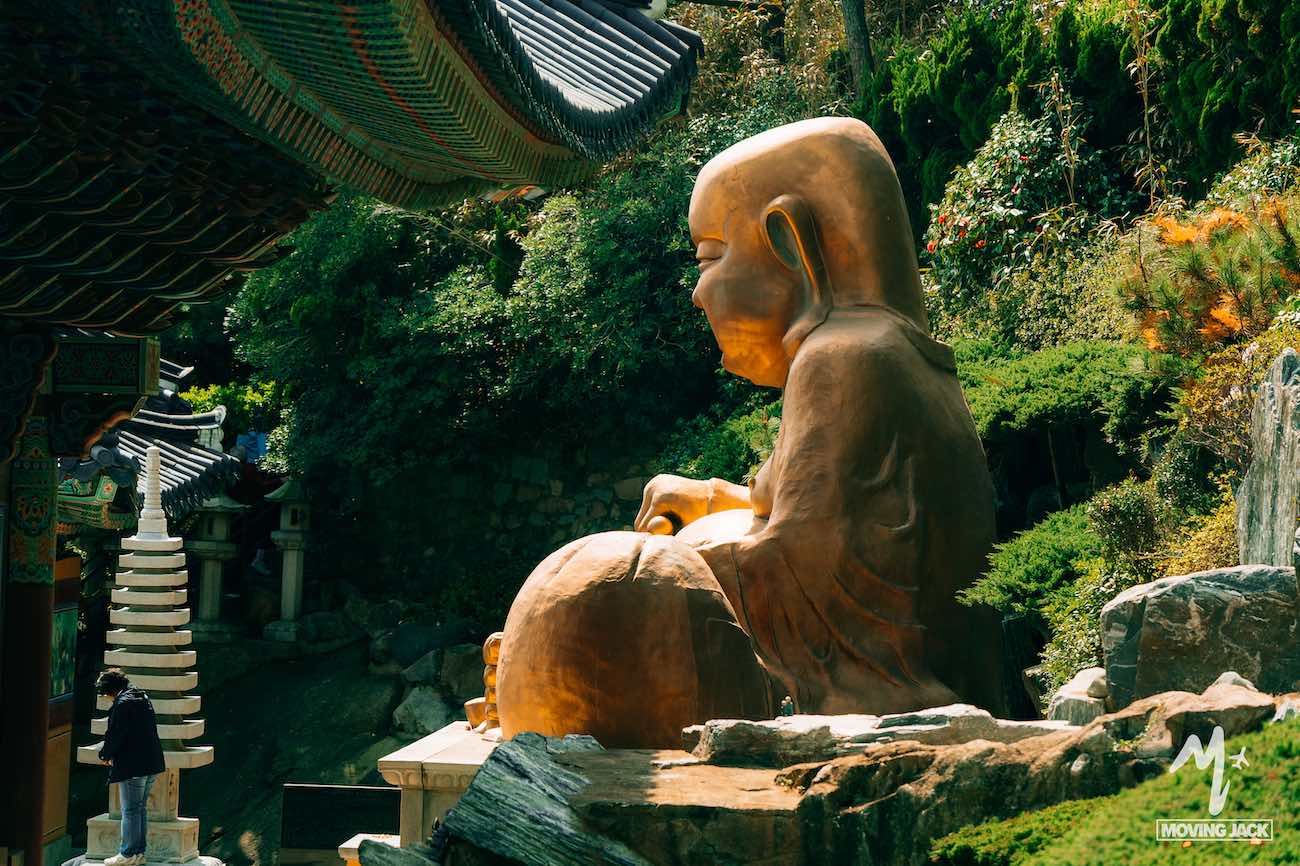 A large golden Buddha statue sits outdoors among greenery, with a person and temple structures nearby—just one reason why many ask, "Is Busan worth visiting?" The serene atmosphere suggests it certainly is.