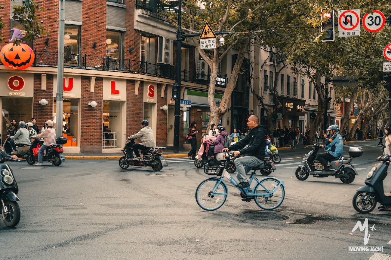 People ride bicycles and scooters through a city street intersection with shops, traffic signs, and a large pumpkin decoration overhead.